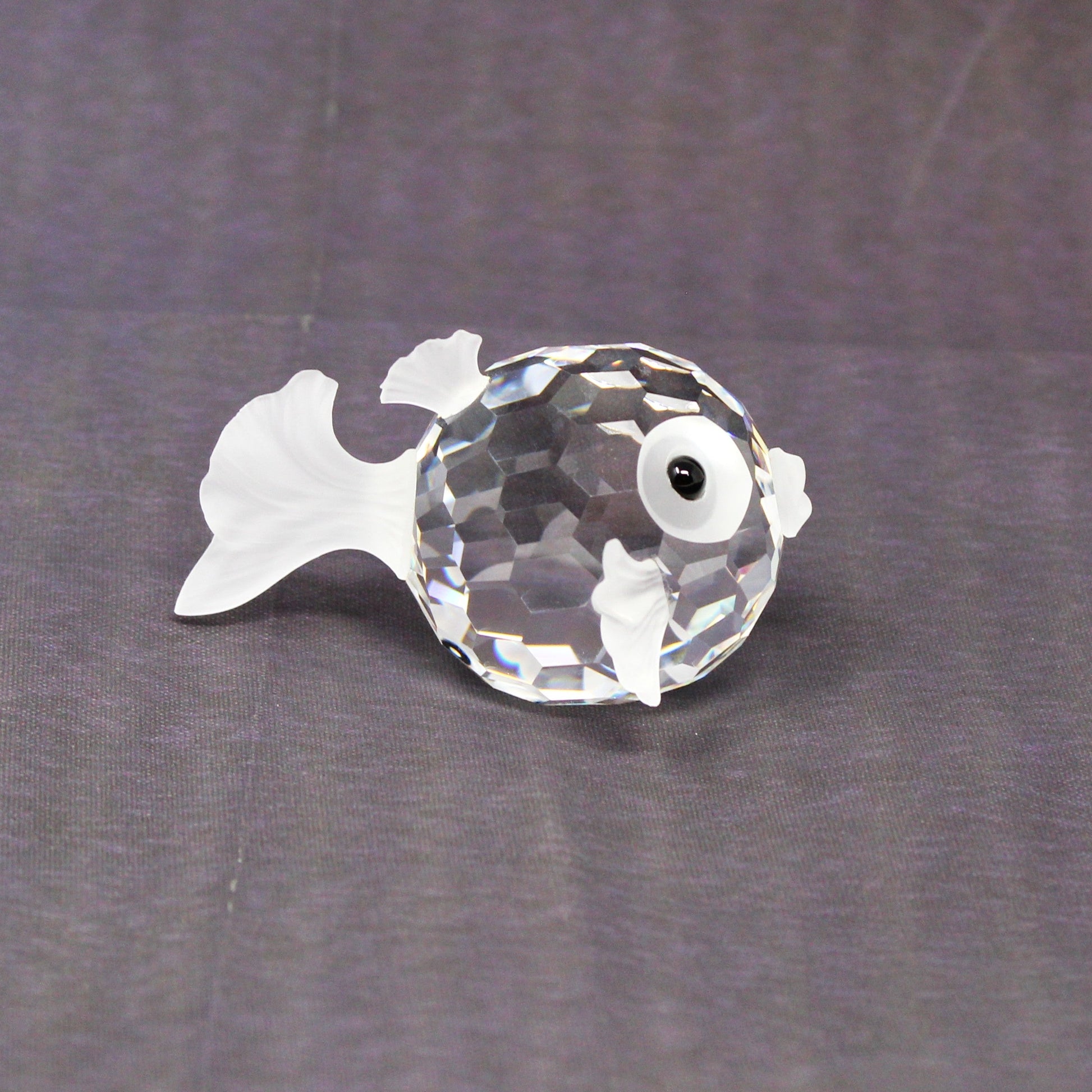 A small clear faceted crystal fish figurine with a white eye, placed on a textured dark gray fabric background.