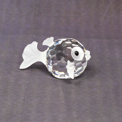 A small clear faceted crystal fish figurine with a white eye, placed on a textured dark gray fabric background.