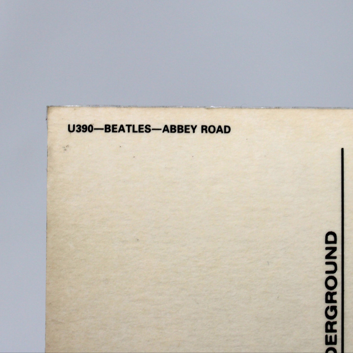 Close-up of the lower-left corner of a beige postcard or printed card with black text reading 'U390-BEATLES-ABBEY ROAD' and postcard lines