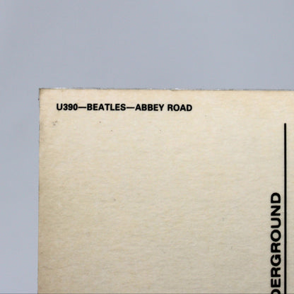 Close-up of the lower-left corner of a beige postcard or printed card with black text reading 'U390-BEATLES-ABBEY ROAD' and postcard lines