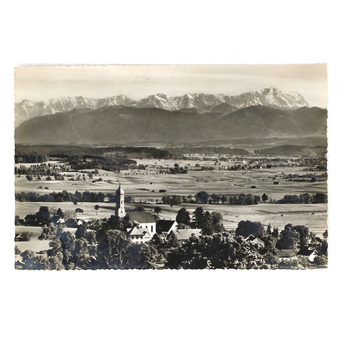 A vintage black and white landscape postcard with real photograph by Perutz film.  Featuring a town with a church steeple in the foreground and a snow-capped mountain range in the background.