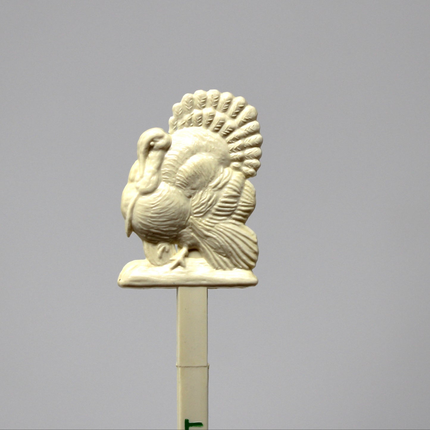 An off-white ceramic or resin turkey topper on a stick, photographed against a solid gray studio background.