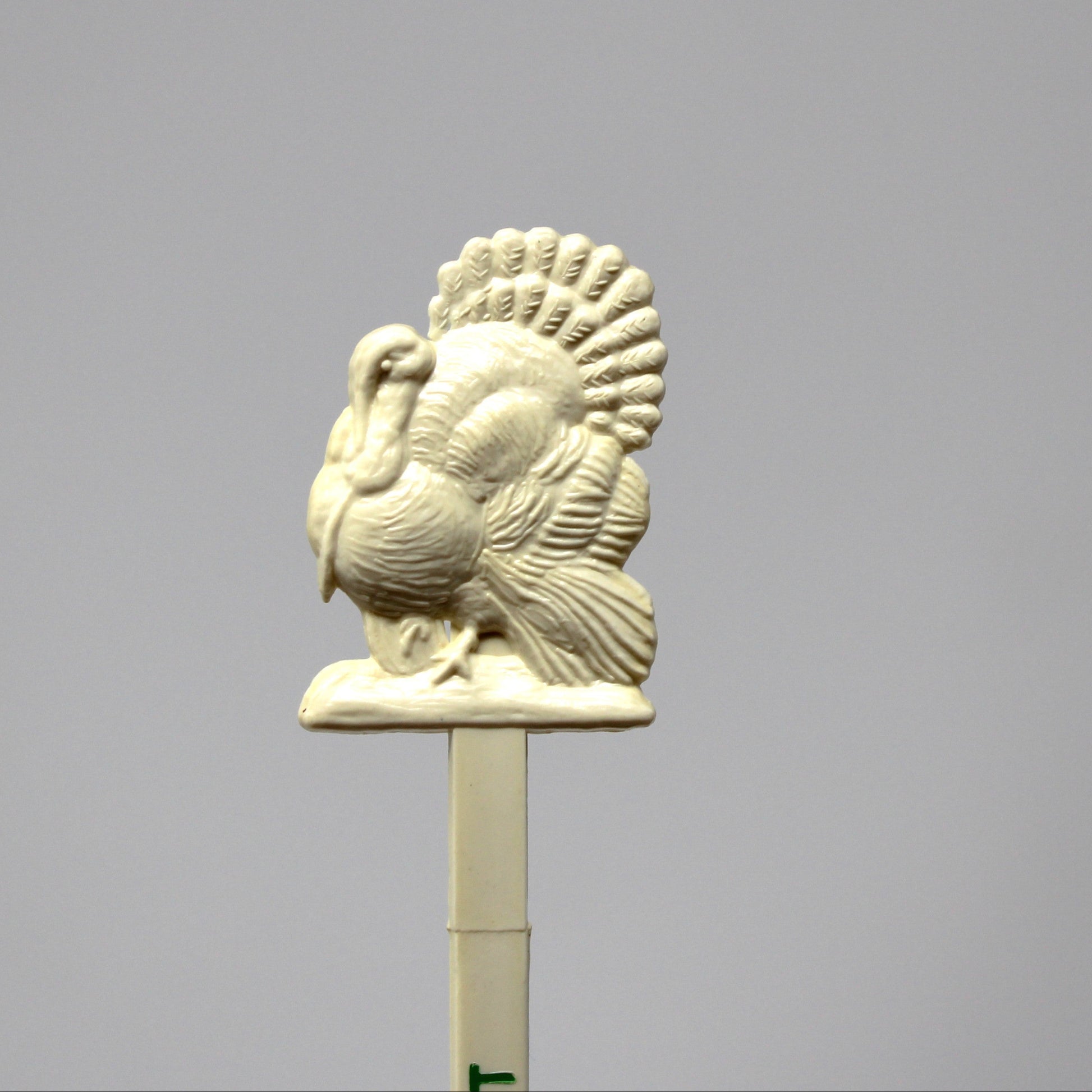 An off-white ceramic or resin turkey topper on a stick, photographed against a solid gray studio background.