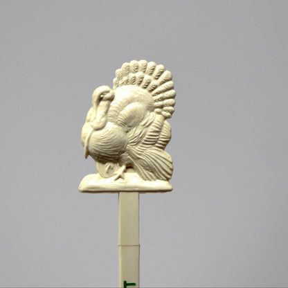 An off-white ceramic or resin turkey topper on a stick, photographed against a solid gray studio background.