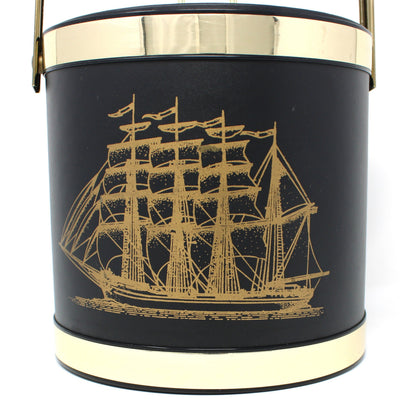 Close up of a vintage ice bucket, black cylindrical bucket-style with a gold-toned handle and trim, featuring a gold ship illustration on the front