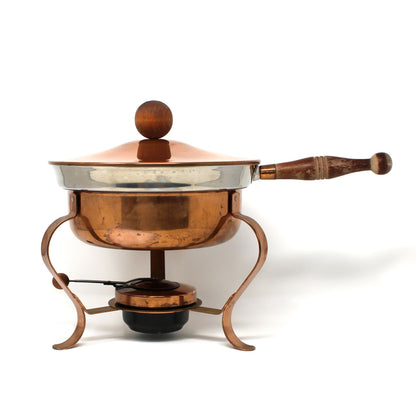A vintage copper chafing dish with a wooden handle and lid knob, resting on a matching copper tripod burner with a small black fuel pan