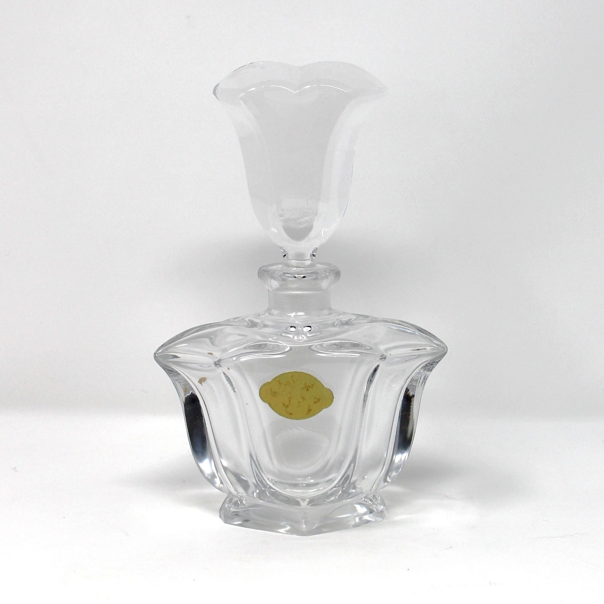 A clear glass decanter with a faceted, ribbed base and a flared stopper, shown on a plain white studio background with a small gold sticker on the front.