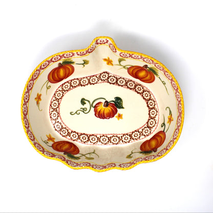 A decorative ceramic pumpkin-shaped serving dish with a hand-painted autumn design featuring pumpkins, leaves, and floral borders, shown on a white background with another dish partially visible on the right.
