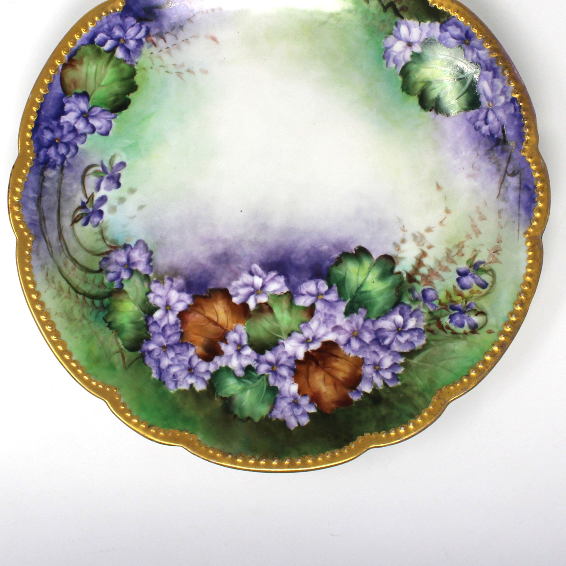 close up view of an antique scalloped Limoges France porcelain plate with a hand-painted floral design featuring purple flowers, green & brown leaves, and a gold moriage rim.