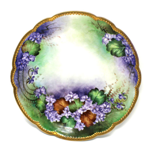 An antique scalloped Limoges France porcelain plate with a hand-painted floral design featuring purple flowers, green & brown leaves, and a gold moriage rim.