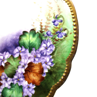A close-up of an antique ornate French Limoges decorative plate with a scalloped gold rim, featuring painted purple flowers, green leaves, and orange-brown accents leaves