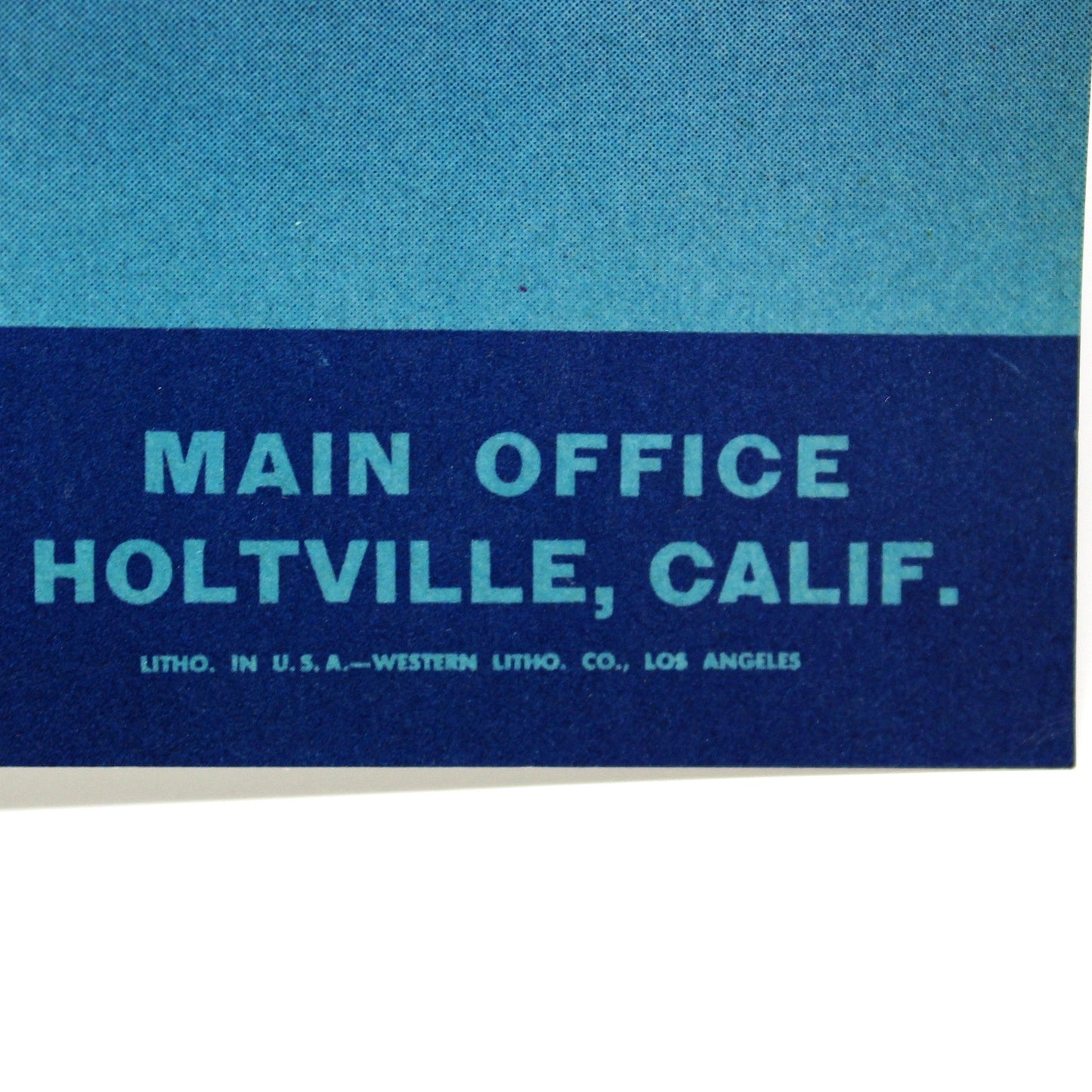 A close-up of a vintage printed sign or poster showing part of a yellow logo at the top and the text 'MAIN OFFICE HOLTVILLE, CALIF.' on a blue background, photographed against a white surface.