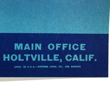 A close-up of a vintage printed sign or poster showing part of a yellow logo at the top and the text 'MAIN OFFICE HOLTVILLE, CALIF.' on a blue background, photographed against a white surface.
