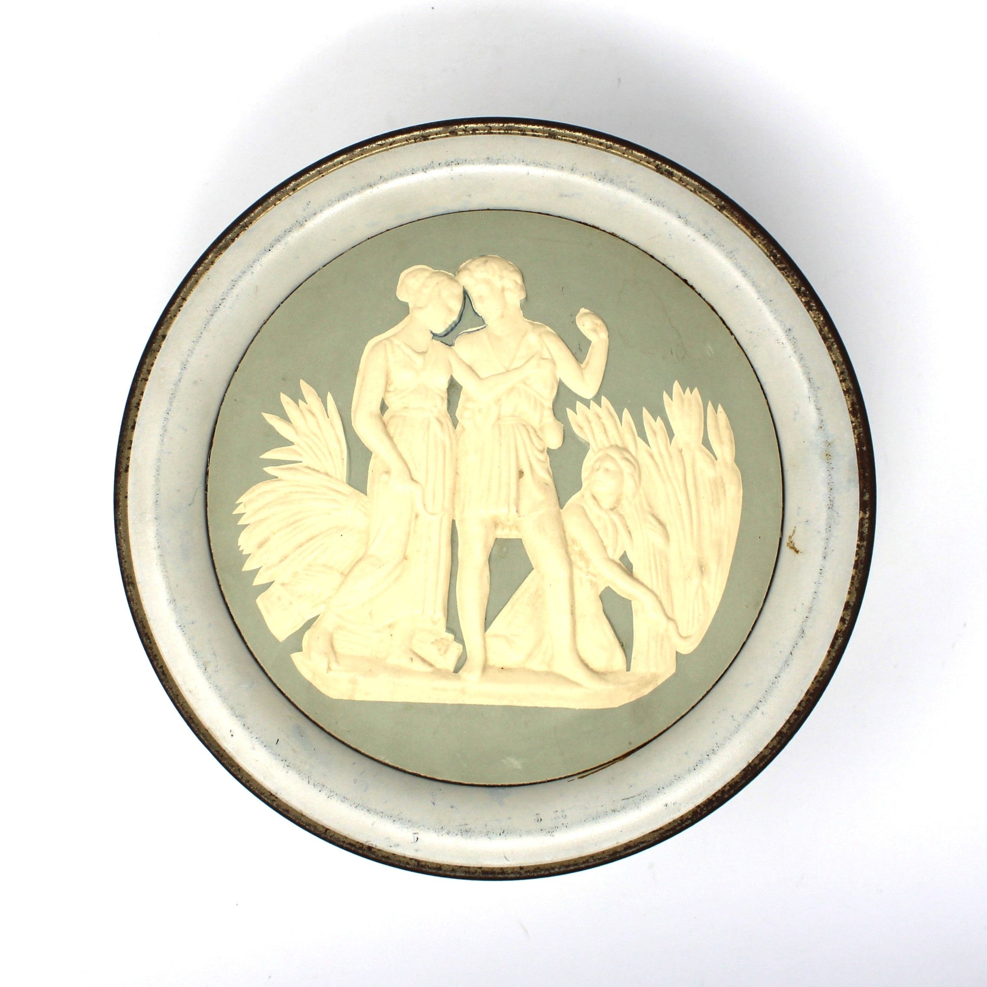 A vintage, round Wedgwood-style storage tin, featuring a pale sage green background with a cream-colored classical relief sculpture of two figures, set against a plain white background.