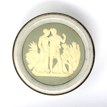 A vintage, round Wedgwood-style storage tin, featuring a pale sage green background with a cream-colored classical relief sculpture of two figures, set against a plain white background.