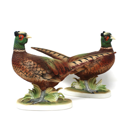A pair of vintage pheasant figurines. The detailed hand-painted porcelain birds feature brown bodies, green necks, and red facial accents, standing on small leafy bases.