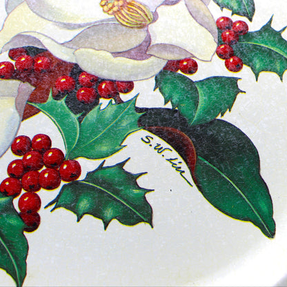 close up of artist signature on A vintage rectangular Christmas serving tray by Potpourri, with a cream-colored background, decorated with a border of white magnolia flowers, green holly leaves, and red berries.