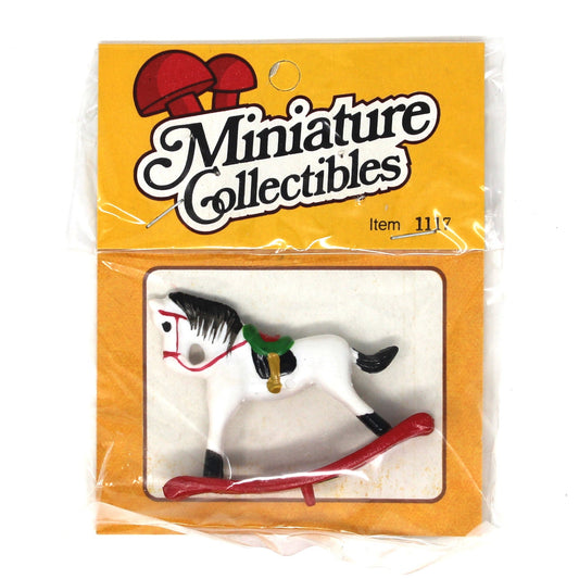 A miniature collectible rocking horse toy in its original packaging. The white plastic toy horse features black and red details, a green saddle, and rests on a red rocking base, all sealed inside a clear plastic bag with a yellow 'Miniature Collectibles' header card.