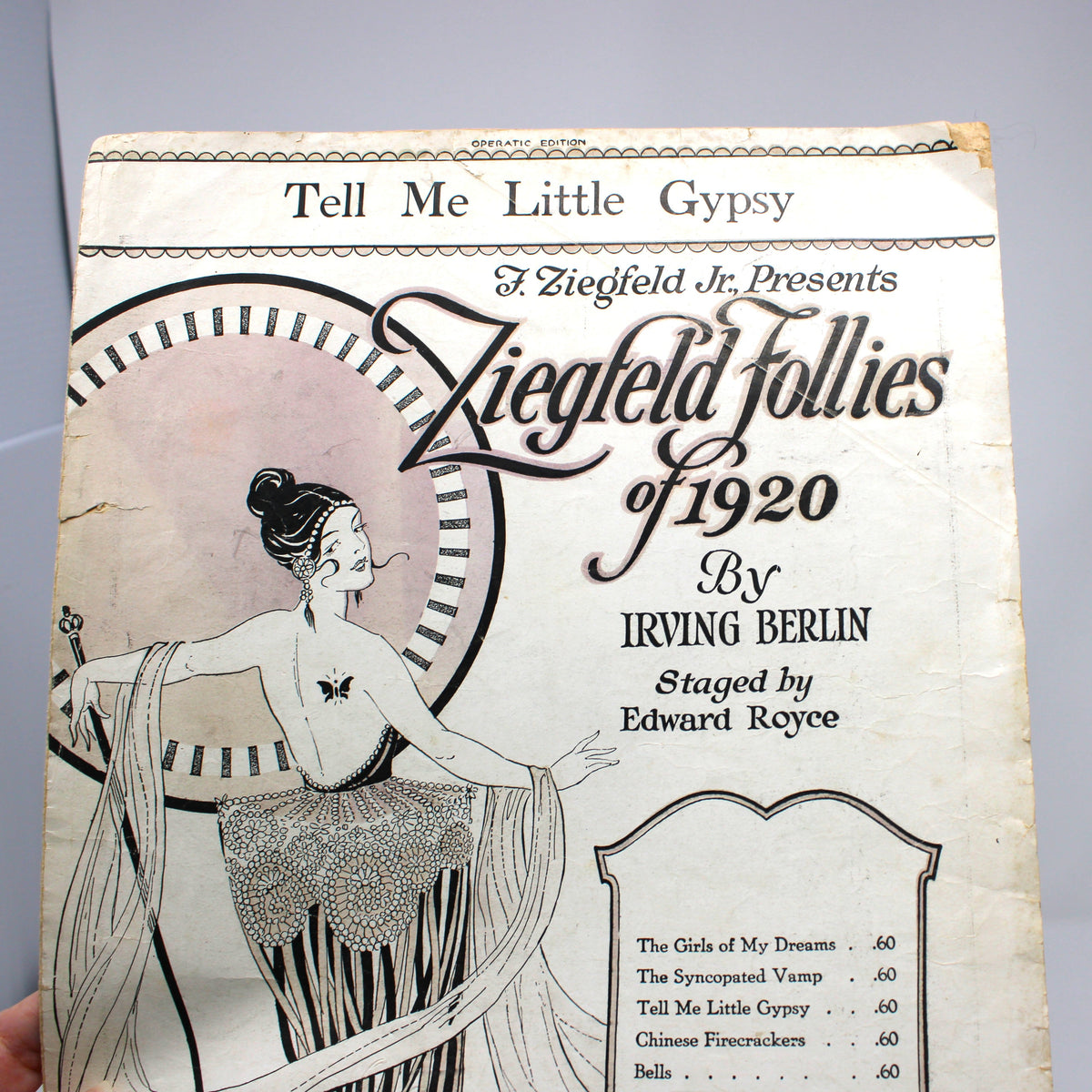 A vintage printed program or booklet cover for 'Ziegfeld Follies of 1920' held by a hand against a plain light gray background. The cover features an Art Deco style illustration of a woman and text including 'Tell Me Little Gypsy' and 'A Ziegfeld Je Presents'.