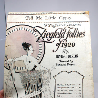 A vintage printed program or booklet cover for 'Ziegfeld Follies of 1920' held by a hand against a plain light gray background. The cover features an Art Deco style illustration of a woman and text including 'Tell Me Little Gypsy' and 'A Ziegfeld Je Presents'.