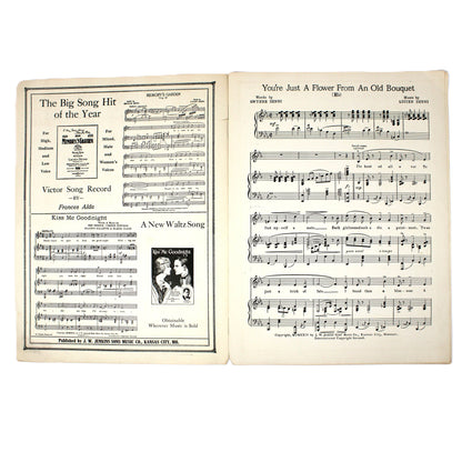 An open vintage sheet music book on a white background, showing printed musical pages with song lyrics and musical notation. 