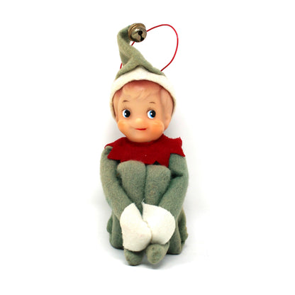 A small elf doll ornament with a green hat, red scarf, and white boots, hanging by a red string on a plain white background.