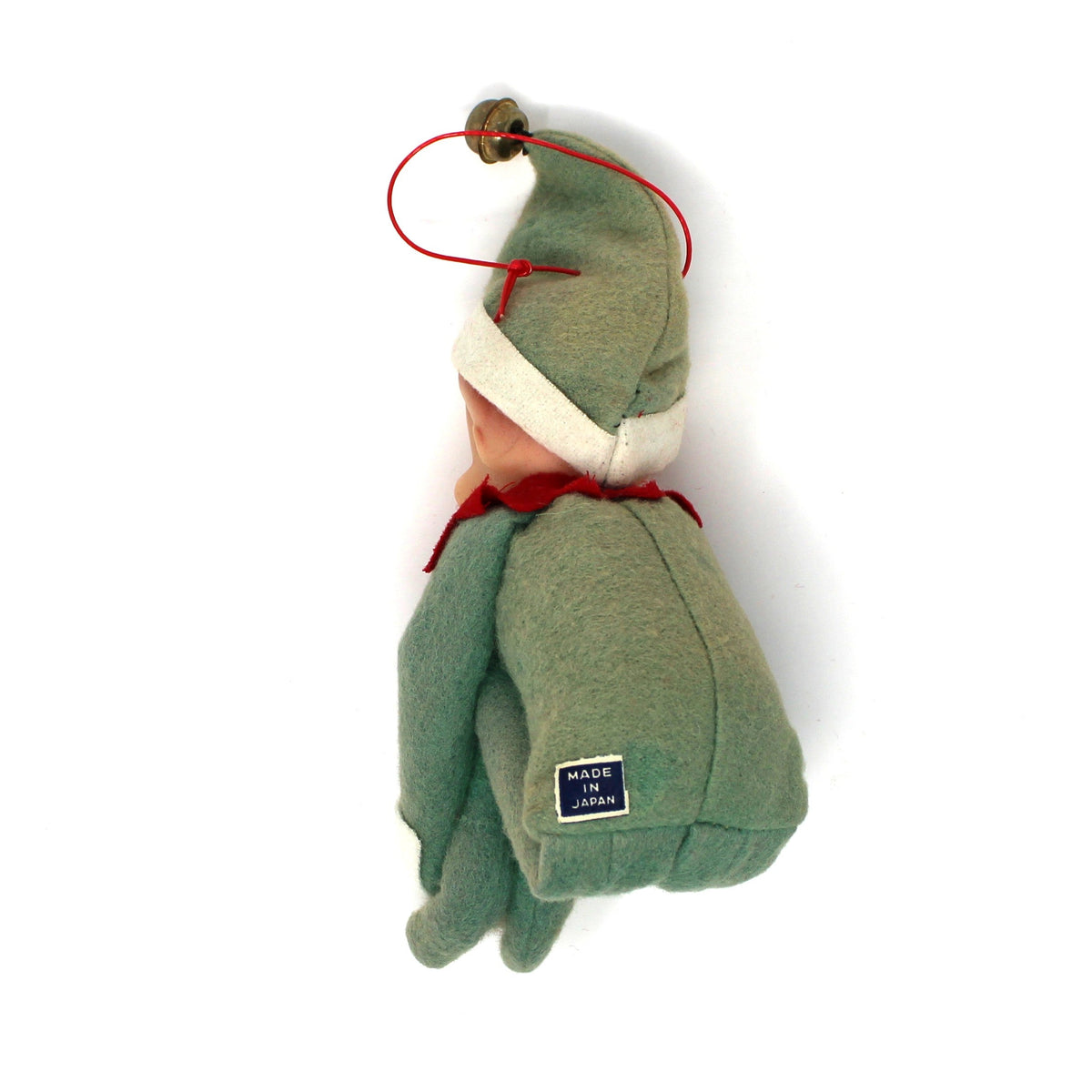 A small handmade plush ornament shaped like a seated figure in muted green, with a cream collar, red cord accents, and a small blue tag near the bottom, photographed on a plain white background.