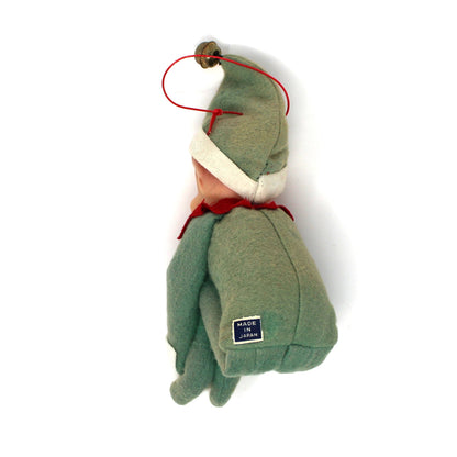 A small handmade plush ornament shaped like a seated figure in muted green, with a cream collar, red cord accents, and a small blue tag near the bottom, photographed on a plain white background.