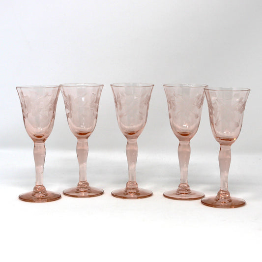 Five small pink tinted glass goblets with stems and circular bases, arranged in a row on a white studio background.
