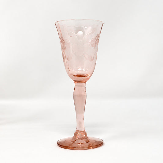 A small pink glass goblet with a textured pattern, standing on a white surface against a light gray background. Another similar goblet is partially visible on the left edge.