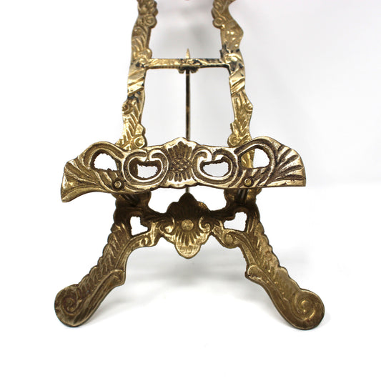 close up view of an ornate antique-style brass easel stand with decorative scrollwork and three legs