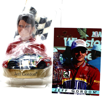 A NASCAR-themed ornament featuring Jeff Gordon in a red racing suit with a checkered flag and gold car base, shown next to a printed collectible trade card with the same driver's image 