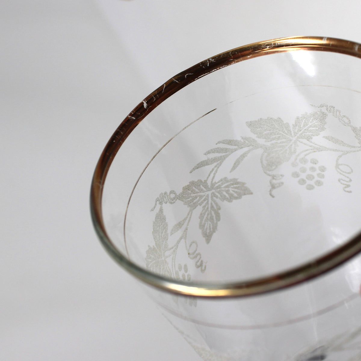 A close-up of a clear glass tumbler with etched floral vine patterns and a metallic gold rim, held by a hand against a plain white background.
